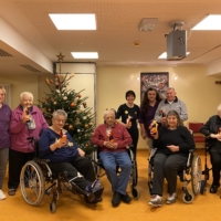 Unsere Mitarbeitenden sind auf einem Gruppenbild mit vier Seniorinnen, zwei Senioren und zwei Pflegerinnen. Ein paar Senior:innen sind in Rollstülen, andere auf einem Rollator. Es hängt ein Kreuz im Eck und einem Christbaum auf der linken Seite des Raumes.