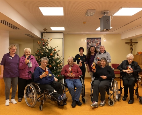 Unsere Mitarbeitenden sind auf einem Gruppenbild mit vier Seniorinnen, zwei Senioren und zwei Pflegerinnen. Ein paar Senior:innen sind in Rollstülen, andere auf einem Rollator. Es hängt ein Kreuz im Eck und einem Christbaum auf der linken Seite des Raumes.