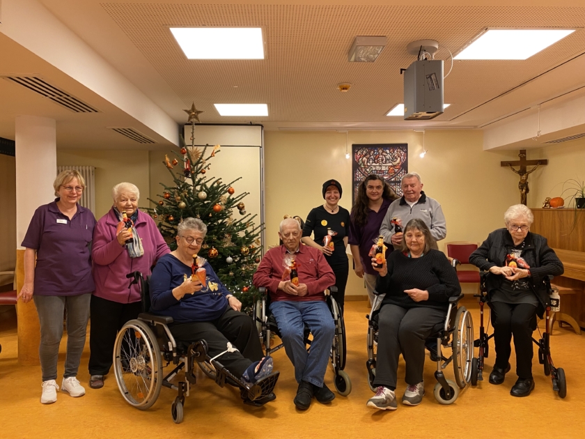 Unsere Mitarbeitenden sind auf einem Gruppenbild mit vier Seniorinnen, zwei Senioren und zwei Pflegerinnen. Ein paar Senior:innen sind in Rollstülen, andere auf einem Rollator. Es hängt ein Kreuz im Eck und einem Christbaum auf der linken Seite des Raumes.