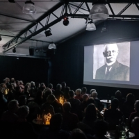 Ein voller, dunkler Saal mit einer leuchtenden Leinwand. Auf der Leinwand ist ein schwarz-weiß Bild von Gustav Haeberle zu sehen. Auf dem Pakett sind kleine Tische mit Stühlen und indirekter Beleuchtung. Die Zuschauer sind von hinten Fotografiert, so dass keine Gesichter zu erkennen sind.