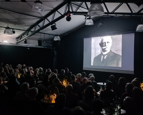 Ein voller, dunkler Saal mit einer leuchtenden Leinwand. Auf der Leinwand ist ein schwarz-weiß Bild von Gustav Haeberle zu sehen. Auf dem Pakett sind kleine Tische mit Stühlen und indirekter Beleuchtung. Die Zuschauer sind von hinten Fotografiert, so dass keine Gesichter zu erkennen sind.