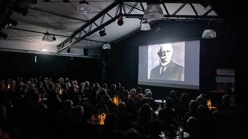 Ein voller, dunkler Saal mit einer leuchtenden Leinwand. Auf der Leinwand ist ein schwarz-weiß Bild von Gustav Haeberle zu sehen. Auf dem Pakett sind kleine Tische mit Stühlen und indirekter Beleuchtung. Die Zuschauer sind von hinten Fotografiert, so dass keine Gesichter zu erkennen sind.